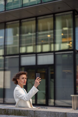 Attractive woman in white suit standing near business centre and making photo. City walking concept