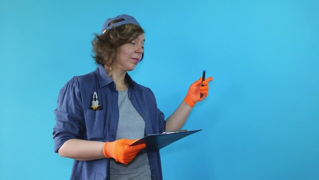 An adult female foreman in a cap and orange gloves holds a paper tablet on a blue isolated background. The woman thinks. Portrait of a woman in a shirt