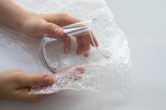 Woman hands packaging a glass for water with white transparent bubble wrap on a white background. Material for packing fragile items for safe transportation. Close-up.