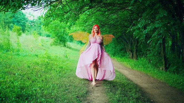 Portrait fantasy woman faerie, butterfly wings costume, lady walks on path summer nature green tree forest. Girl elf fairy tale angel. Pink dress long hem train slow motion. Happy smilig pretty face