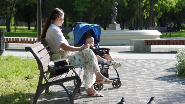 Mother with little cute child in baby stroller in the park, woman is resting on a bench on a sunny day.