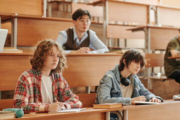 Group of youthful interracial students in casualwear making notes in copybooks while sitting by...