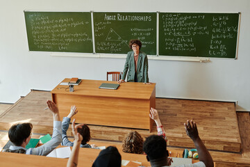 Group of interracial students raising hands to ask questions to teacher standing by blackboard with...