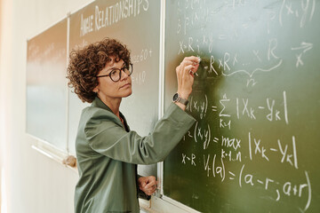 Serious female teacher of algebra with piece of chalk writing down equations on blackboard while...