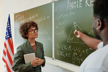 Young serious female teacher of geometry listening to African American student with piece of chalk...