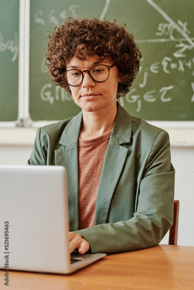 Poster young serious teacher concentrating on work with electronic documents or tables while typing on lapt