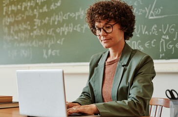 Young confident teacher typing on laptop keypad while sitting by workplace in front of blackboard...