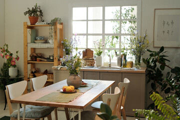 Cozy kitchen with wooden table in the center surrounded by sink with kitchenware and wildflowers by window and green domestic plants