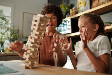 Young woman and her youthful daughter trying to hold on fallen tower built up of wooden blocks while sitting by table during game