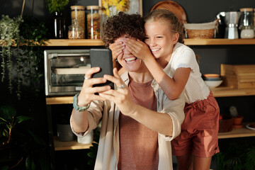 Cute youthful girl covering eyes of her mother with smartphone by hands while having fun during selfie against shelves with kitchenware