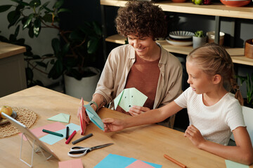 Young woman and youthful girl sitting by table and comparing their handmade origami items after...