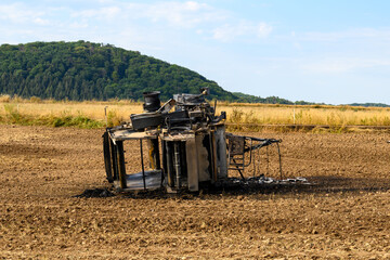 Top view of a burnt out hay baler lying in a field against a green forest