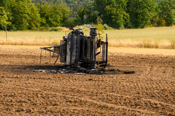 Underside of a burnt out hay baler in a field with trees and grass in background