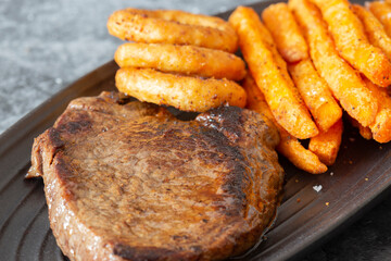 Rump steak dinner, with sweet potato fries and onion rings, on a black oval plate.  On a dark stone background