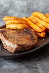 Rump steak dinner, with sweet potato fries and onion rings, on a black oval plate.  On a dark stone background