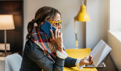 a woman with yellow glasses talks on the phone in her office while reading an agenda . she is...