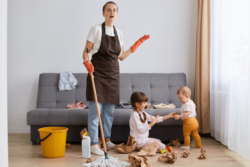 Portrait of tired mother wearing brown apron cleaning at home, woman with mop cleaning with kids...