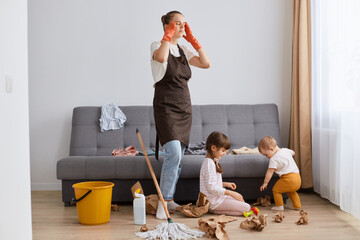 Image of tired mother standing near her daughters, mommy cleaning at home, woman touching head,...