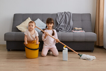 Indoor shot of little girl with mop in hands and her infant sister doing household the living room...