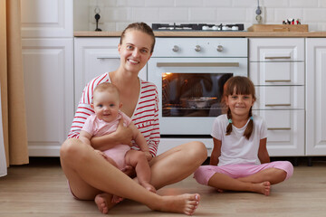 Image of satisfied happy smiling woman wearing striped shirt sitting on floor in kitchen with her little daughters, mother holding infant baby, people looking at camera with optimism.