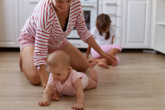 Image of faceless woman wearing striped shirt sitting on floor in kitchen with her little daughters, infant baby crawling, playing with mommy, family spending time at home.