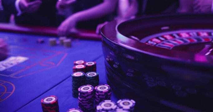Vibrant casino table with roulette in motion, with casino chips, tokens, the hand of croupier, dollar bill money and a group of gambling rich wealthy people playing bet in the background
