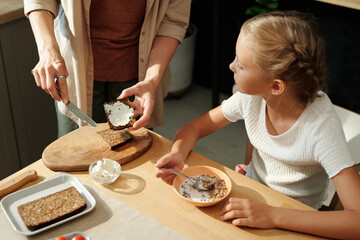 Cute youthful girl eating muesli with milk and looking at her mother making sandwich with dairy...