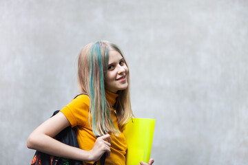 Teen schoolgirl holding backpack and document folder on a grey concrete background. Smiling blond girl with colored hair strands going to lessons. Back to school concept. Copy space.