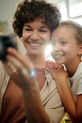 Cheerful young brunette woman with retro photocamera taking photo of herself and her cute youthful daughter standing near by