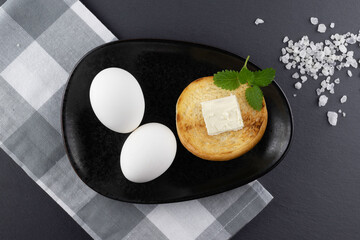 White eggs in a black plate and a slice of hot toast with butter.Breakfast concept.Top view.