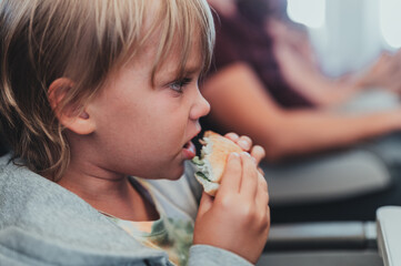 little candid kid boy five years old eats burger or sandwich food sitting in airplane seat on flight traveling from airport. children take a bite. child in air plane eating lunch or dinner meal