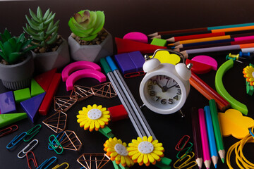 Colorful pencil, pen, clock, paperclip, flowers, plastic geometrical figures for studing on black table. Variety of stationery