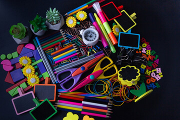 Bright school stationery on black desk. Top view on heap of scissors, small chalkboards, pen, pencil, clock, marker, paperclip, table flowers
