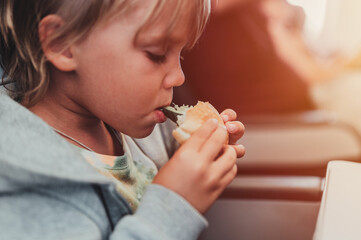 little candid kid boy five years old eats burger or sandwich food sitting in airplane seat on flight traveling from airport. children take a bite. child in air plane eating lunch or dinner meal. flare