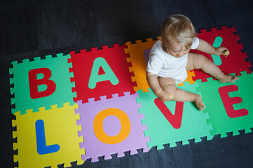 bird view of colorful kids puzzle mat playground in nursery with letters like Baby Love written on them lying on the floor while one year old blond baby in white body is playing
