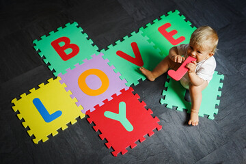 Naklejka premium bird view of colorful kids puzzle mat playground in nursery with letters like Baby Love written on them lying on the floor while one year old blond baby in white body is playing 