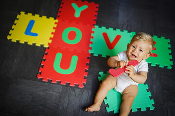 Naklejka premium bird view of colorful kids puzzle mat playground in nursery with letters like Baby Love written on them lying on the floor while one year old blond baby in white body is playing 