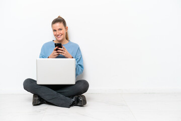 Young woman with a laptop sitting on the floor sending a message with the mobile