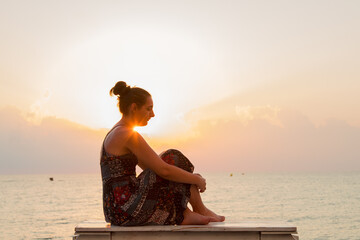 A young woman sits barefoot on the oceanfront bench and waits for the sunrise on a warm summer...