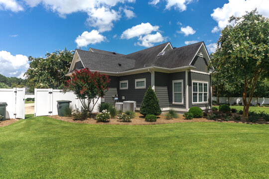 The side view of the backyard of a large gray craftsman new construction house with a landscaped yard and white pvc fence