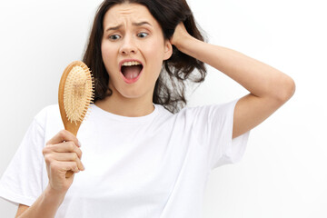 a sad, upset, puzzled woman with long black hair stands in a white T-shirt on a light background and holding her hand on her head in panic looks at her wooden comb