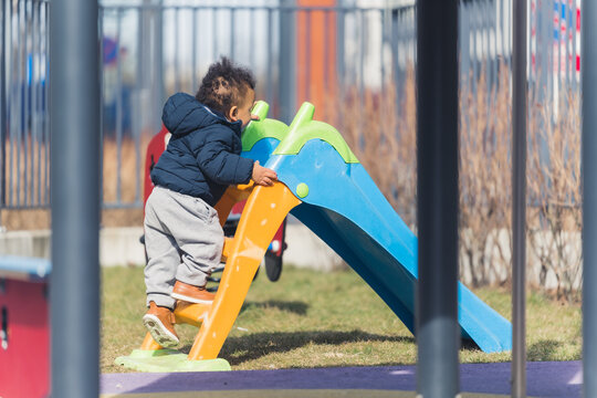 An african ethnicity 5 years old boy dreassed in winter clothing is making his way for a slide inj achildren's playground surrounded by metal fence. High quality photo