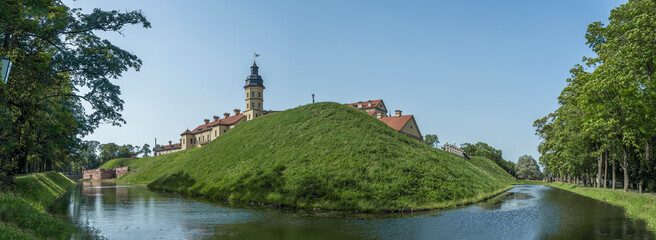 Fototapeta premium Moat of old castle. Moat with reflection in the water and trees. Sunny summer day. Palace, tower, corner towers and moat.