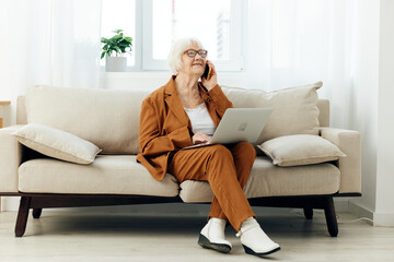Obraz premium an emotional elderly woman with gray hair in a brown stylish suit and white boots is sitting on a beige sofa holding a laptop on her lap working from home