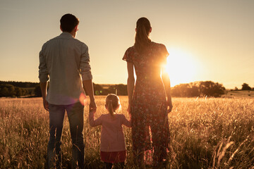 Family of three standing in a meadow holding hands facing the sunset. 