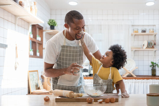 Happy smiling Black son touching his father face with flour while doing bakery at home, Cheerful African American family cooking baking cake or cookie in the kitchen