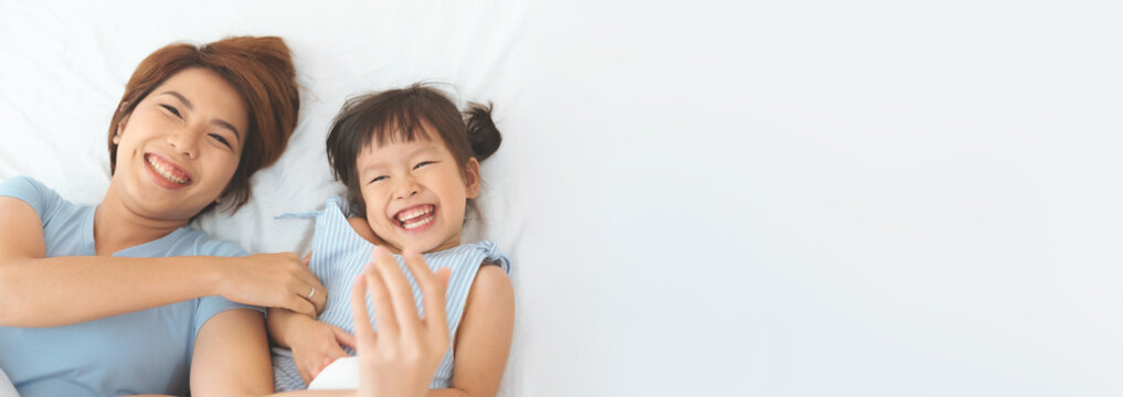 Happy Young asian mother playing ticklish and laughing with her cute daughter on bed indoor, Selective focus at girl, Banner, Copy space