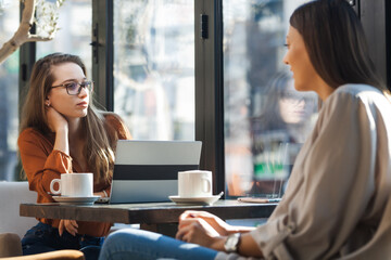 Two young business women in a cafe having one on one meeting. Friends after work talking gossiping and having coffee at a window table on a sunny day..
