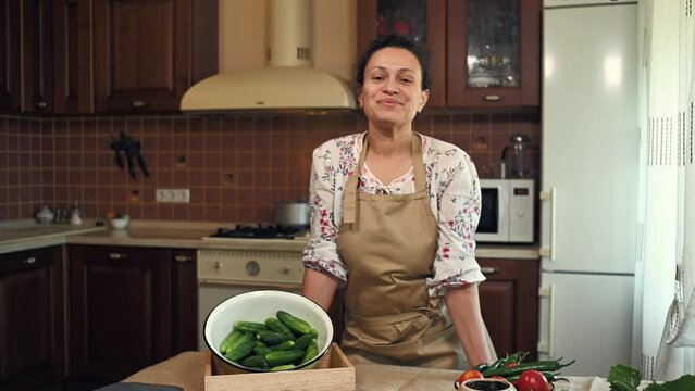Charming woman, housewife, wearing an apron, cutely smiles looking at camera, standing at kitchen table with an enamel bowl of freshly harvested cucumbers and ingredients for pickling at home kitchen