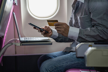 Young businessman holding a smartphone and credit card online working on a laptop sitting on an airplane